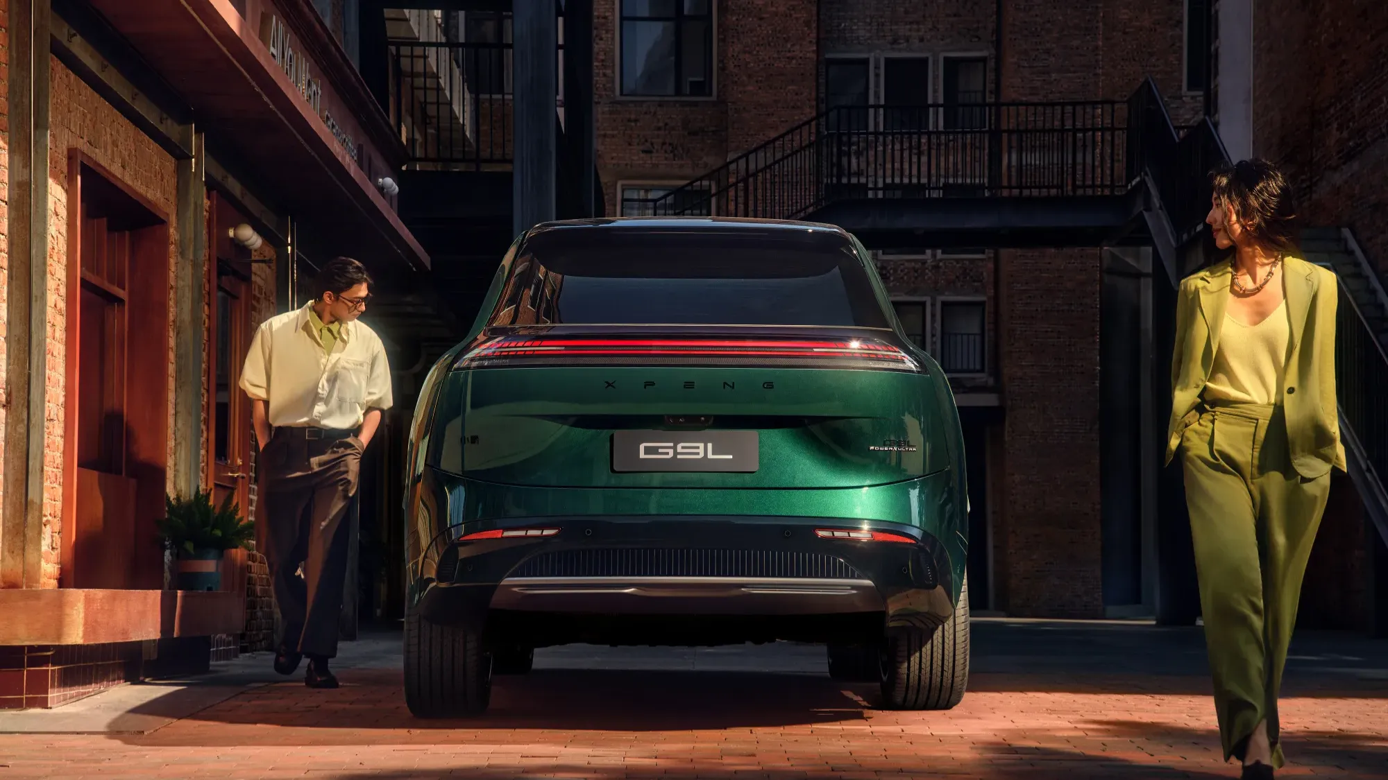 Rear three-quarter view of the green XPeng G9L on a cobblestone street, with two people walking past