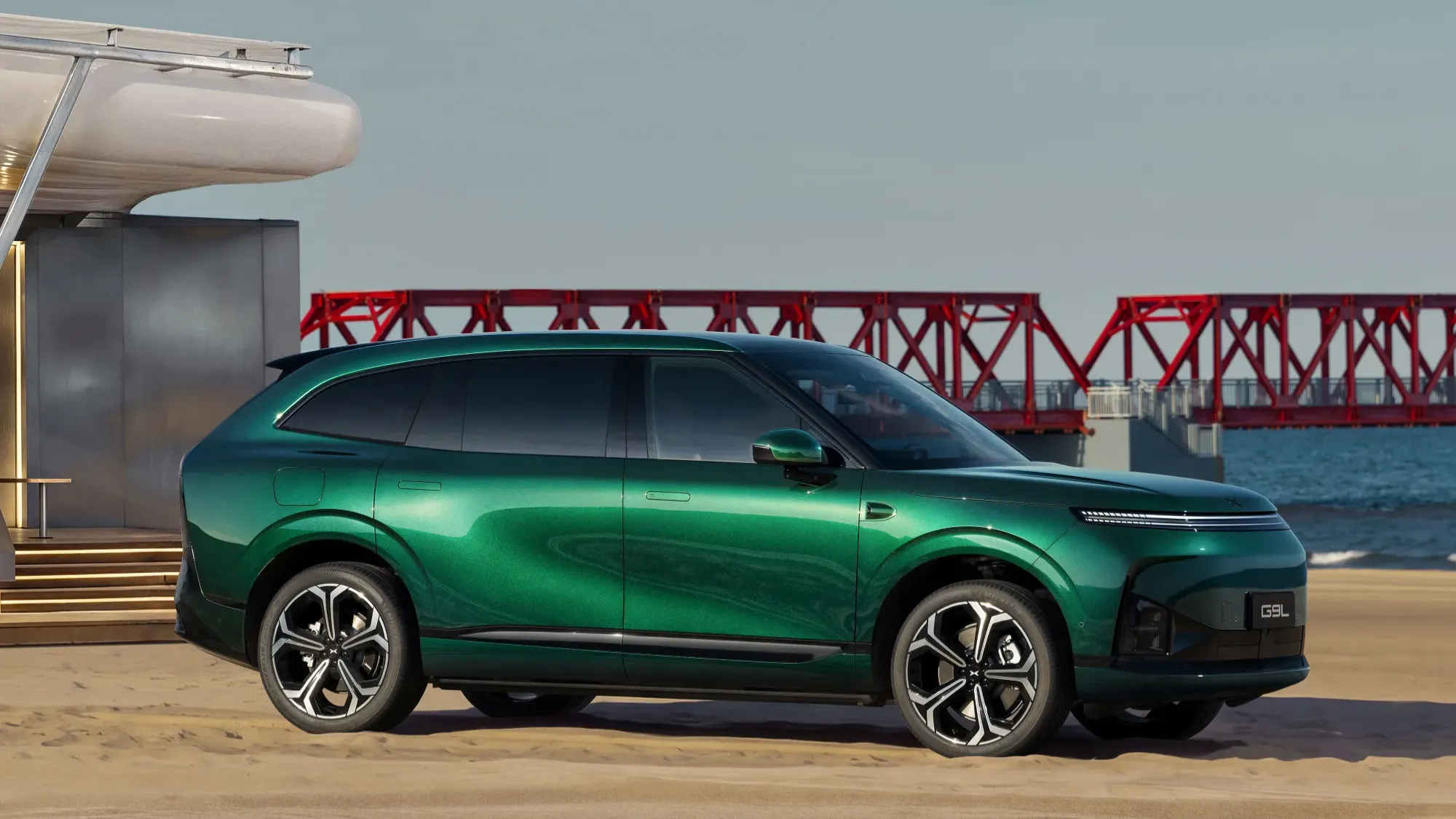 Side profile of the metallic green XPeng G9L SUV parked on a waterfront plaza beside a red truss bridge