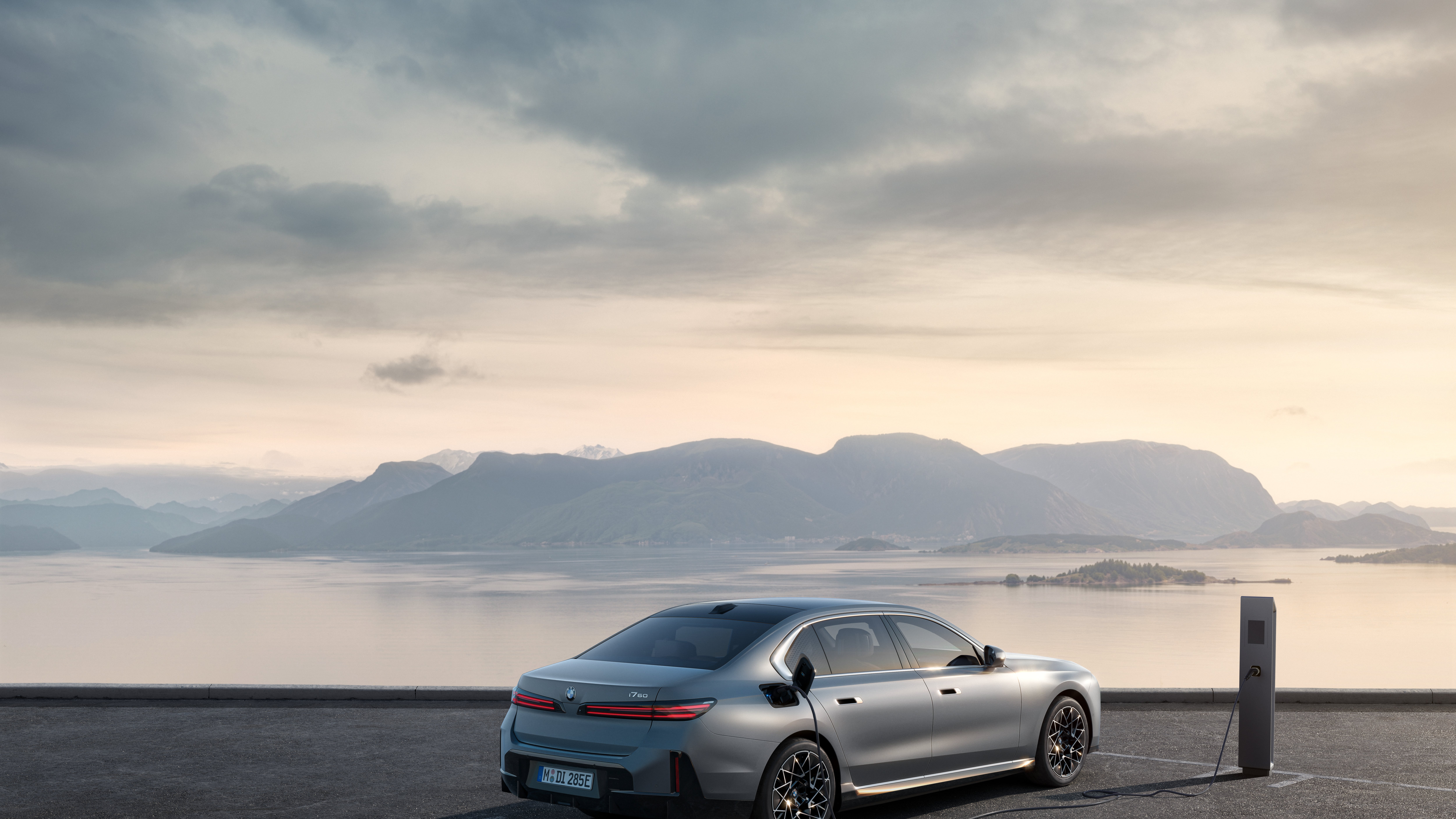 Rear three-quarter view of a BMW i7 60 xDrive plugged into a DC charger beside a fjord at dusk