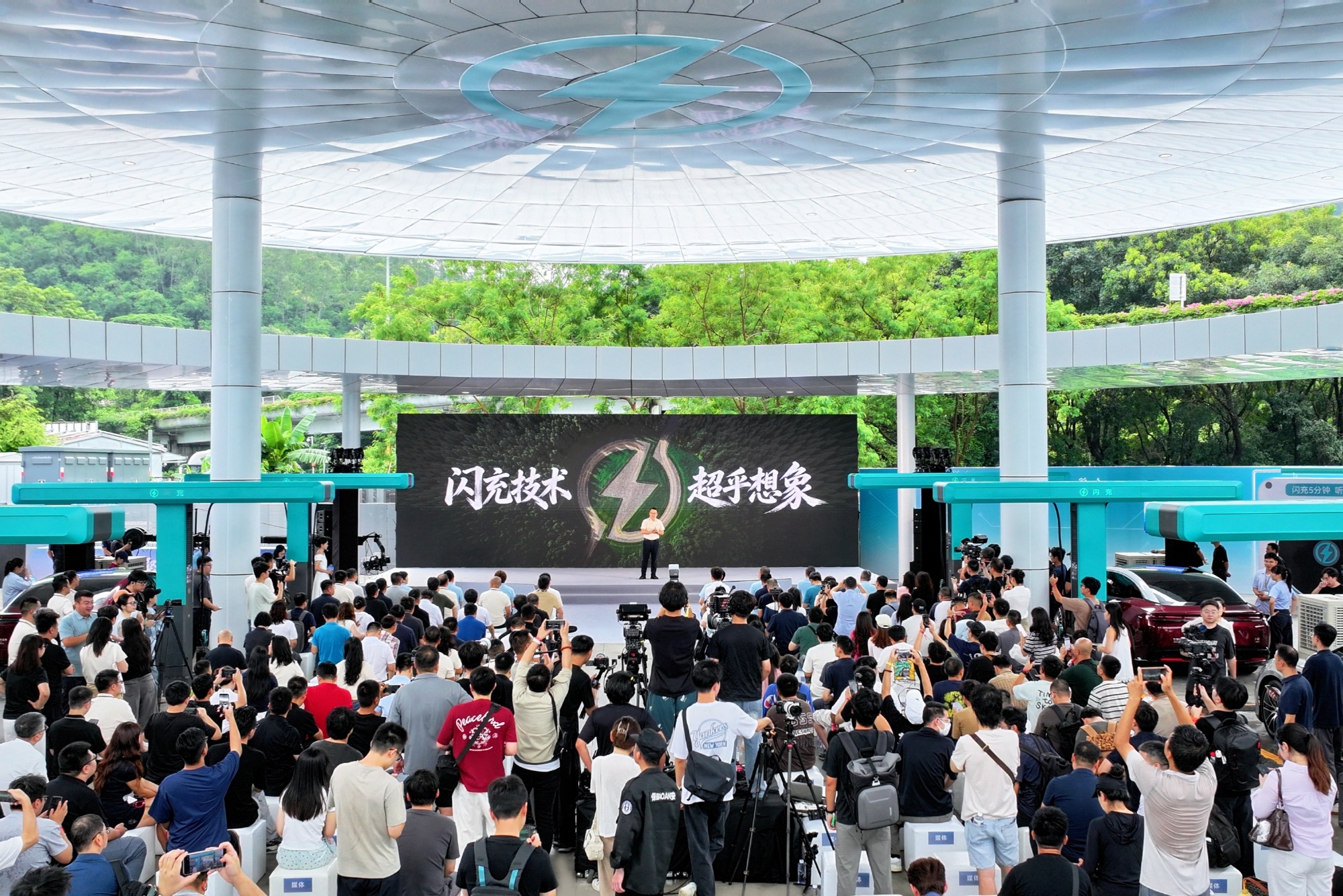 Crowd gathers under the canopy of a BYD flash charging station in Shenzhen as a speaker presents on stage