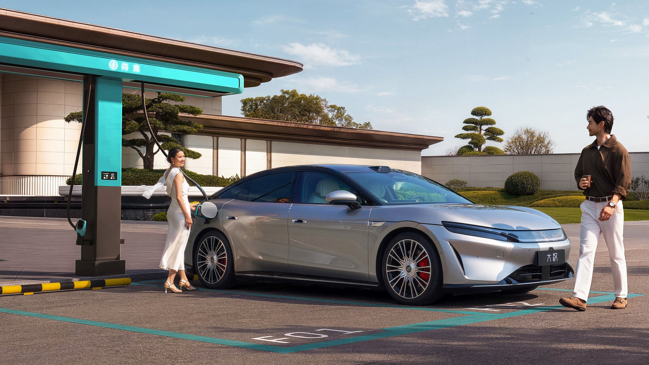 Silver BYD Da Han EV parked in a flash-charging bay with its charge cable plugged in, two people beside the car