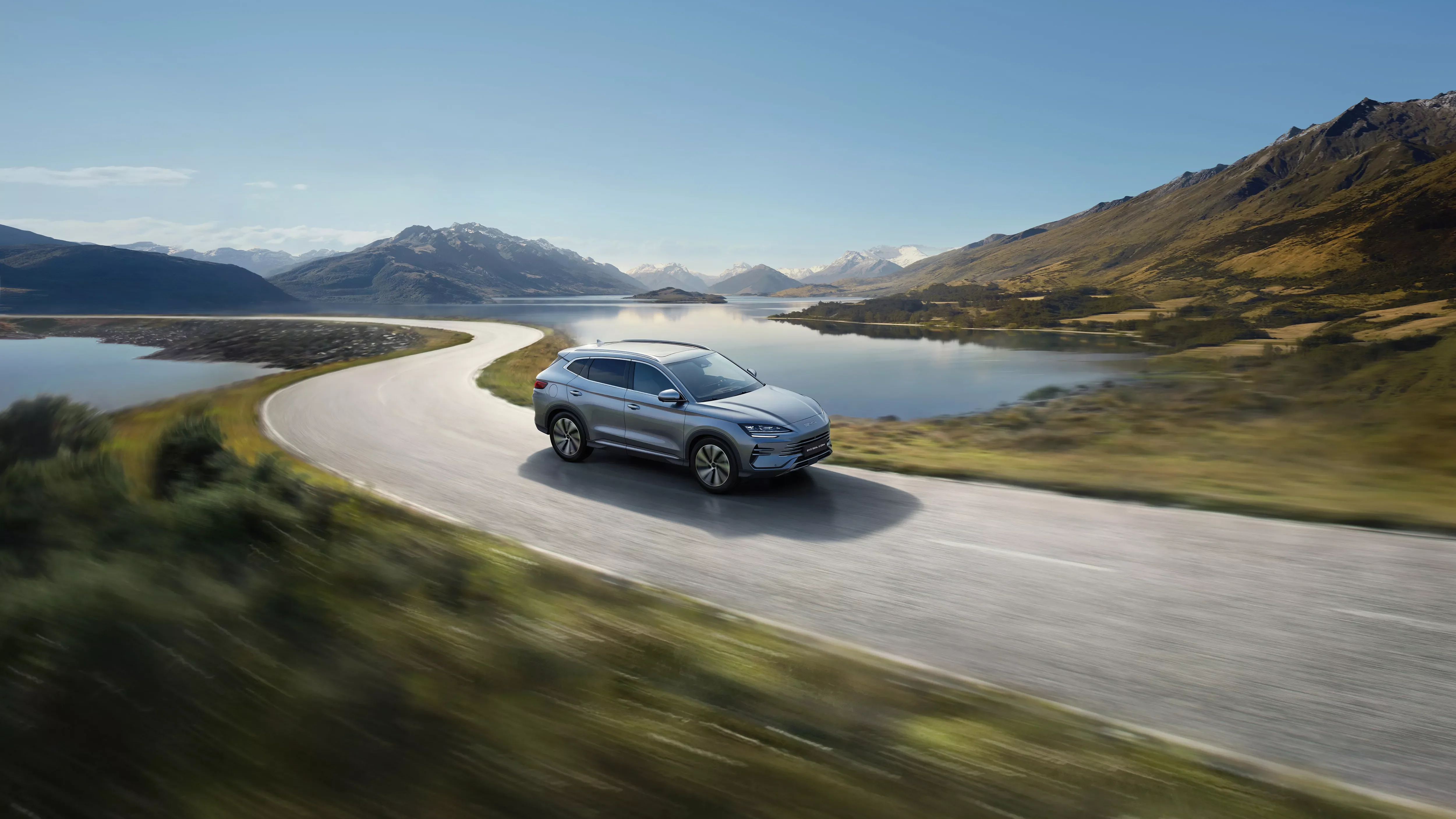 Silver BYD Seal U DM-i driving along a curving lakeside road with mountains behind, from BYD's official imagery