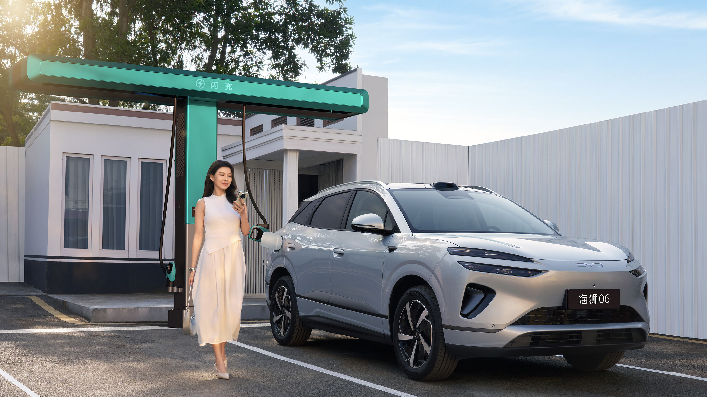 Silver 2026 BYD Sealion 06 EV connected to a flash charger at a BYD station with a woman standing beside it