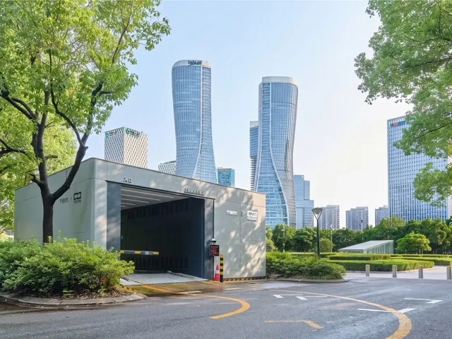 Grey Choco-Swap battery swap station with an open bay in a leafy city plaza, glass skyscrapers behind it