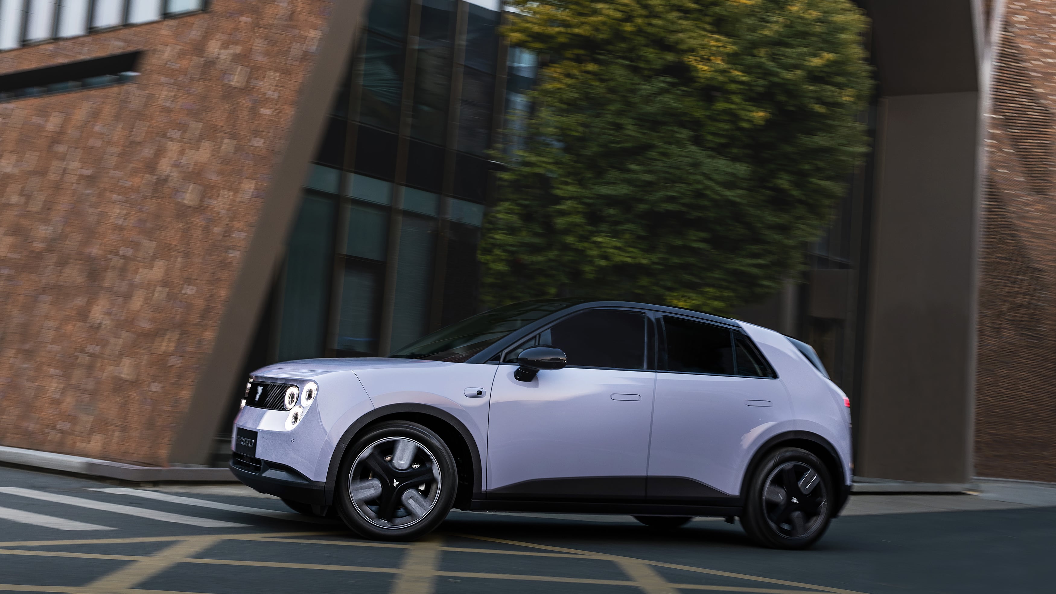 Lavender Firefly hatchback driving past a brick and glass building, seen in side profile with motion blur