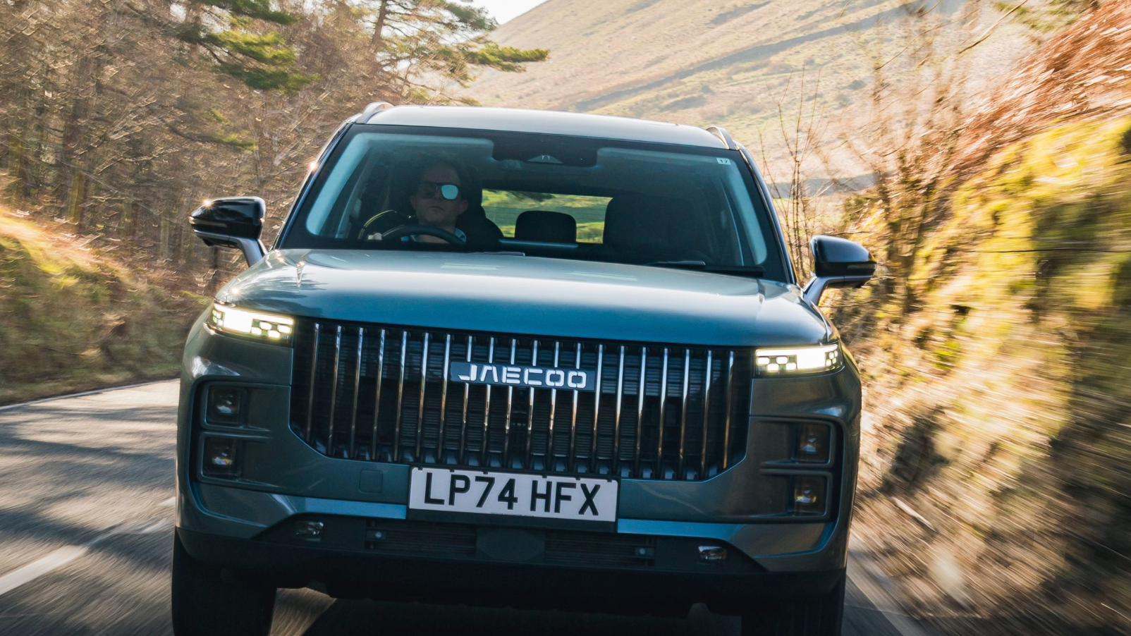 Grey Jaecoo 7 SUV on UK registration plates driving head-on along a sunlit country road with a driver at the wheel
