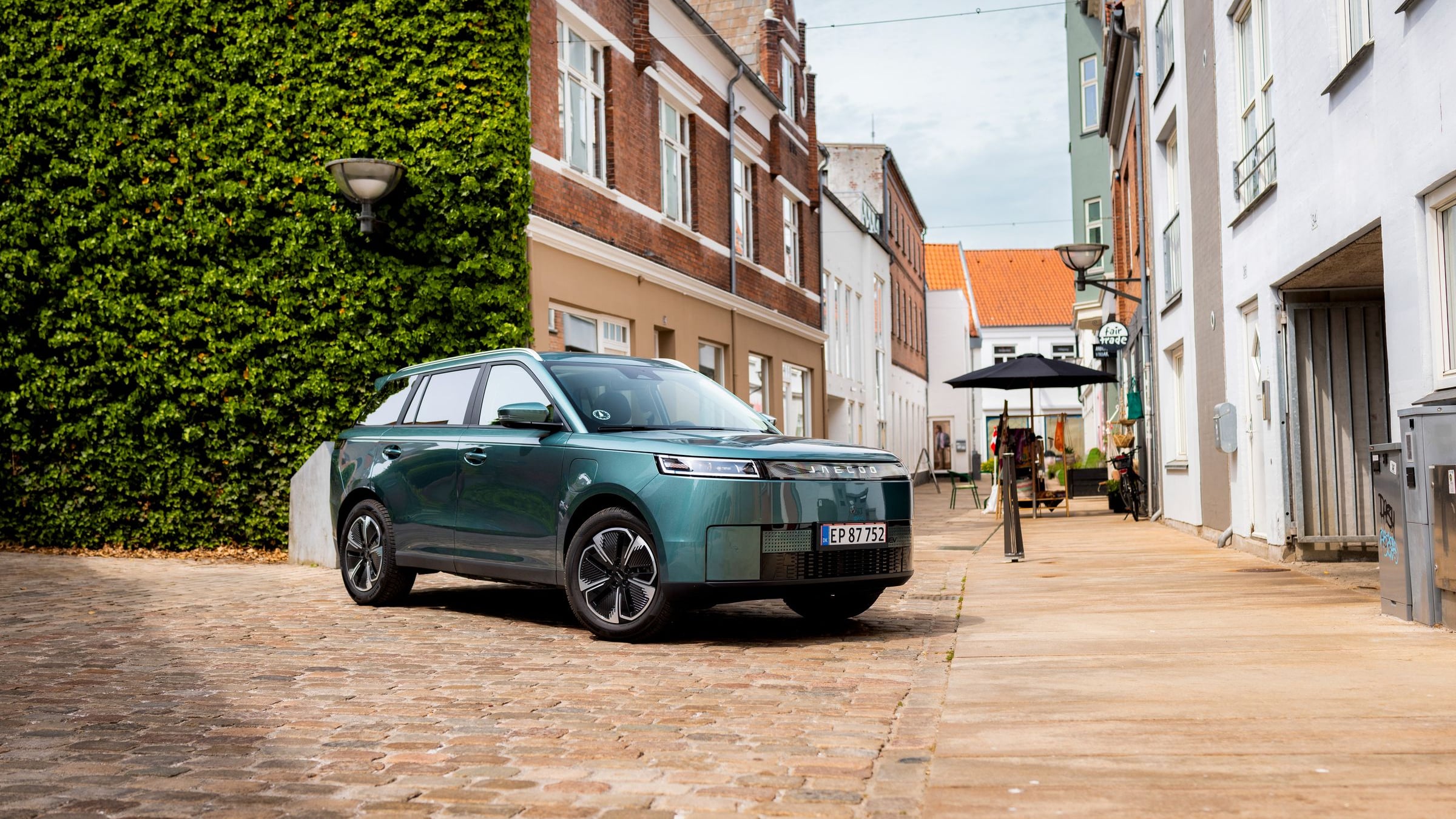 Green Jaecoo J5 EV parked on a cobblestone street lined with brick buildings and an ivy-covered wall in Denmark