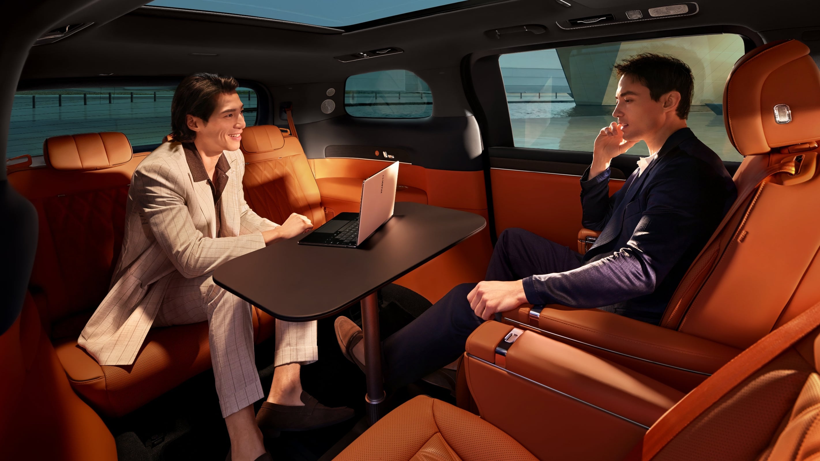 Two passengers face each other across a fold-out table with a laptop in the Luxeed V9 MPV's orange leather rear cabin