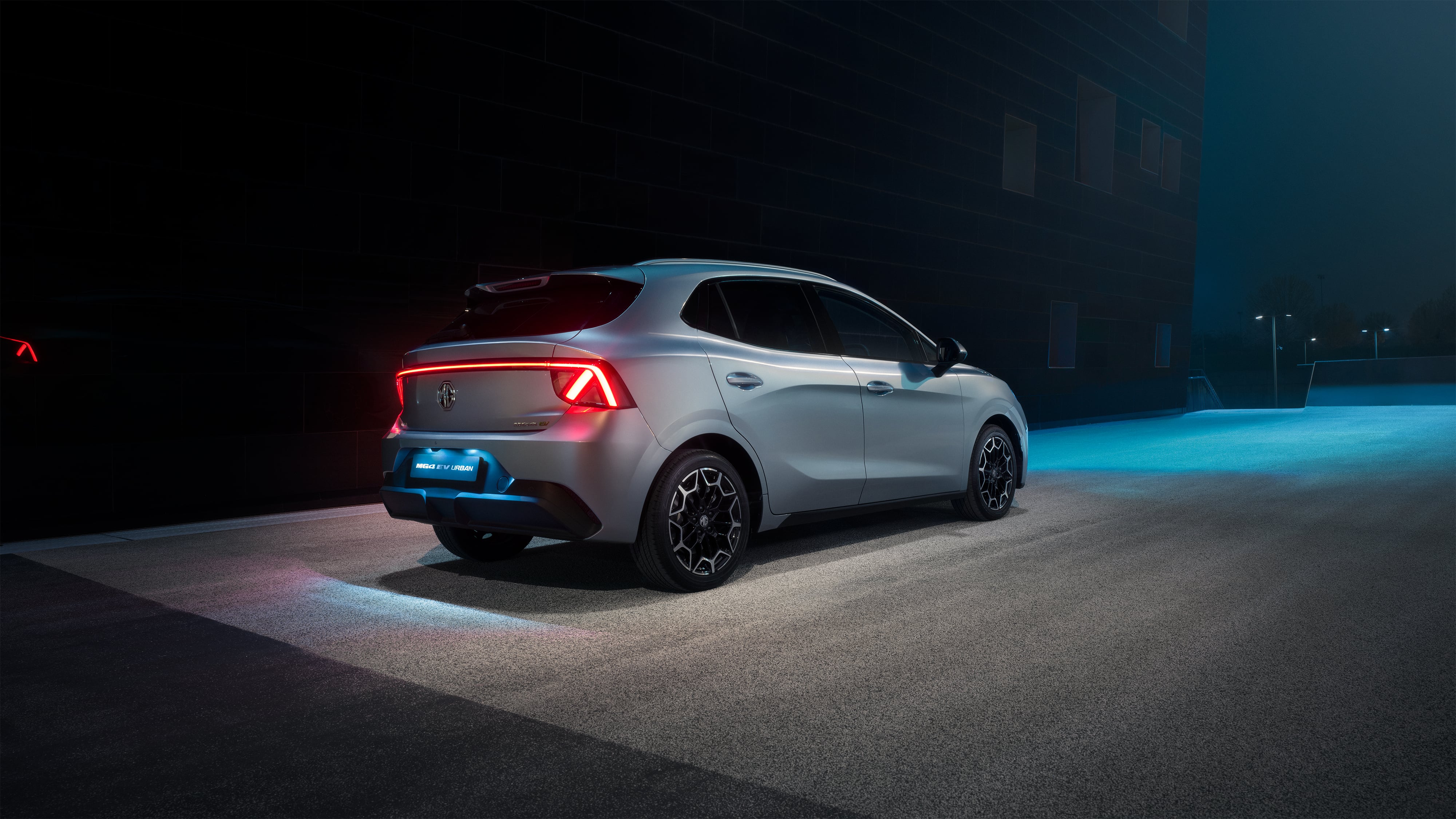 Rear three-quarter view of a silver MG4 Urban at night, showing its full-width LED tail light bar