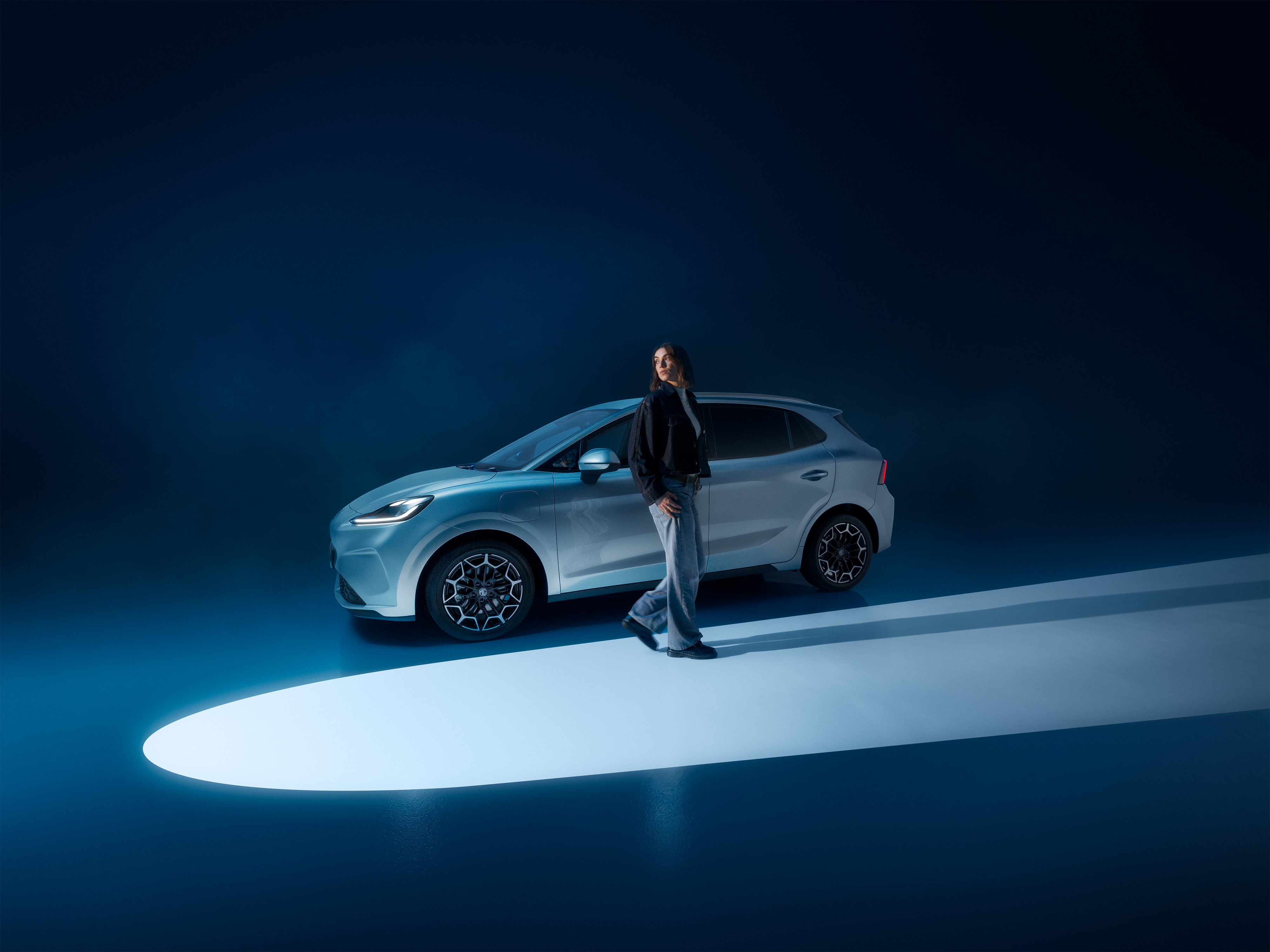 Light blue MG4 Urban in a dark studio with a model walking past it through a spotlight beam