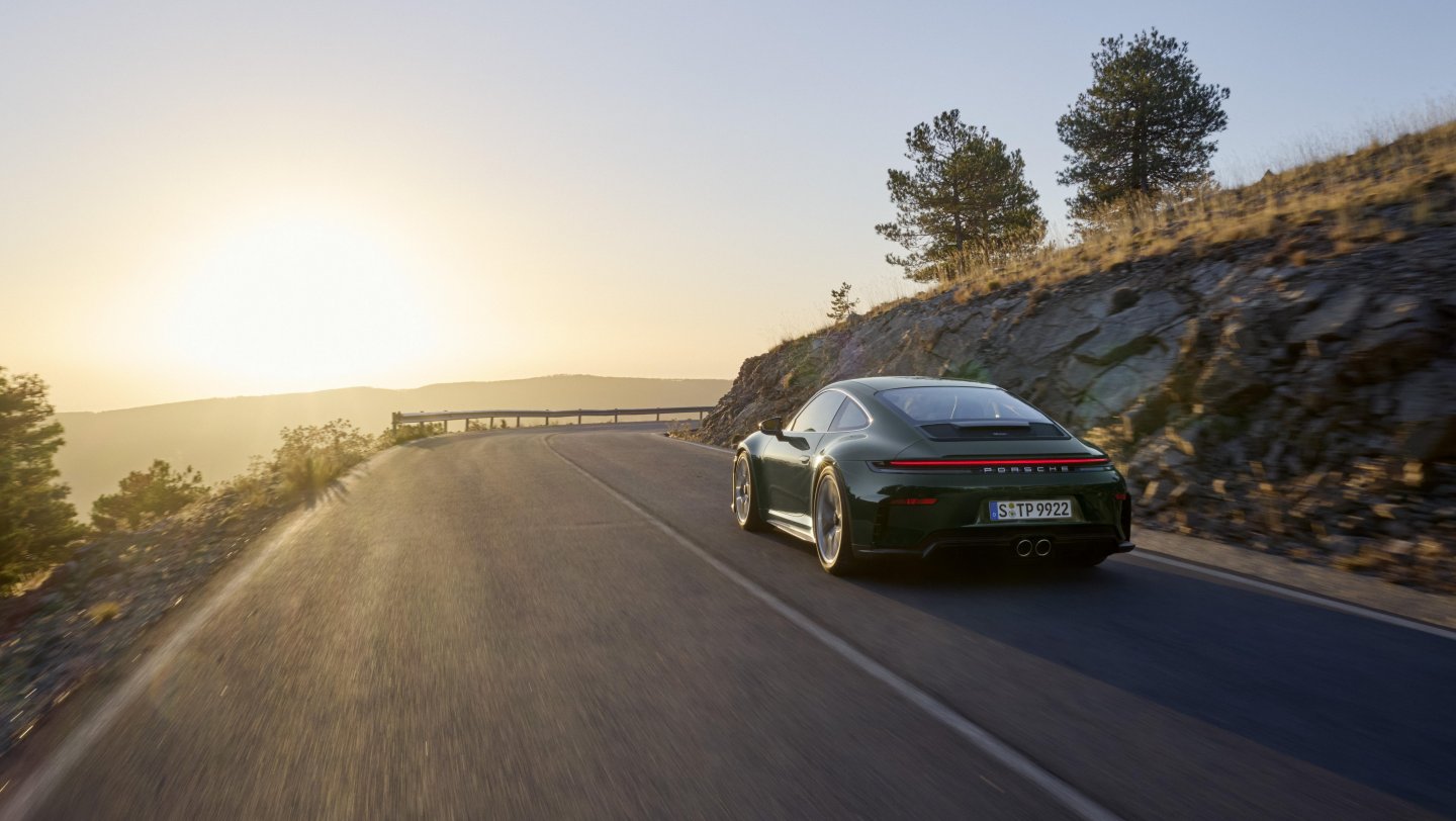 Dark green Porsche 911 GT3 Touring driving on a mountain road at sunset, rear three-quarter view