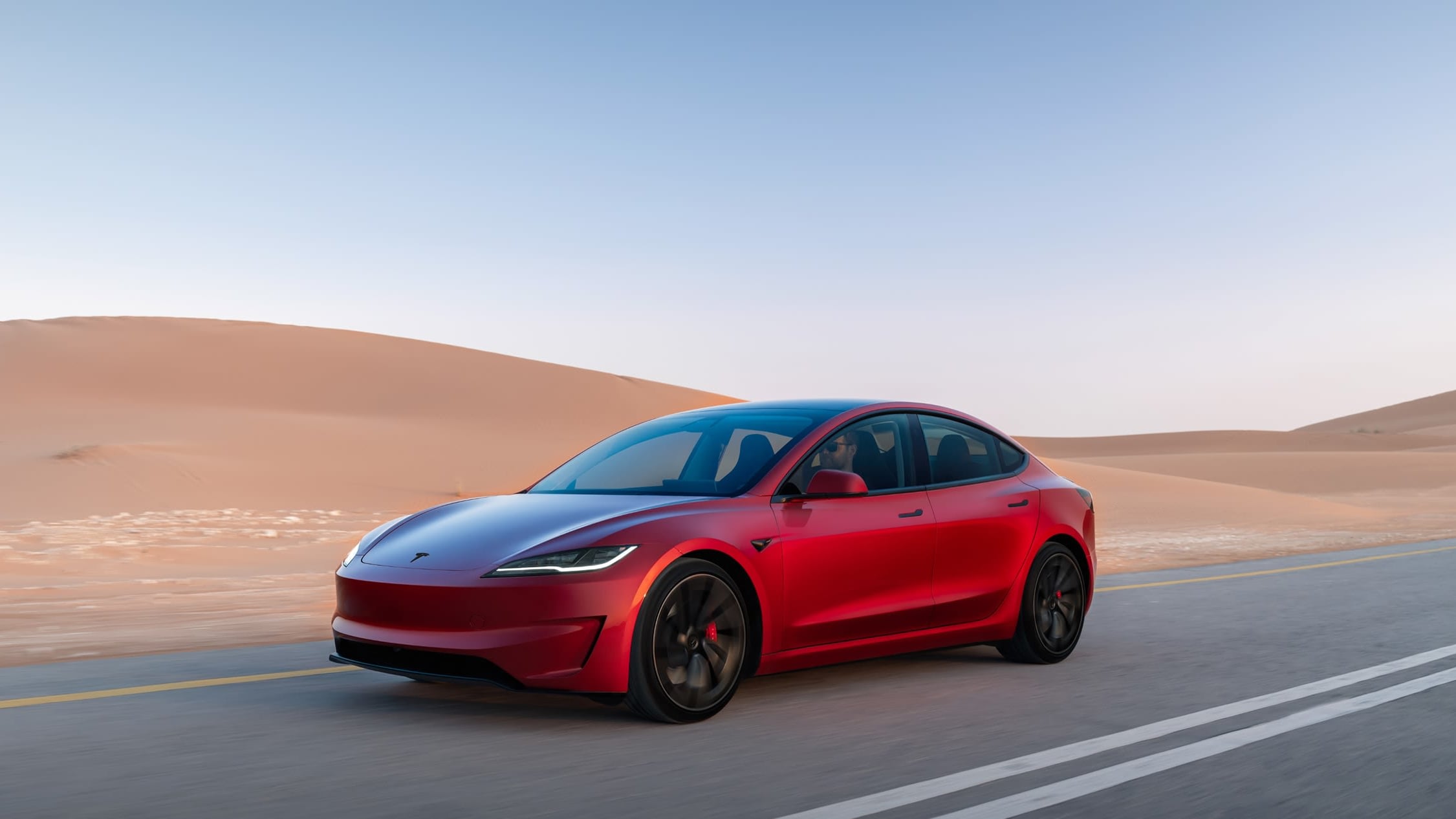 Red Tesla Model 3 Performance with dark wheels and red brake calipers driving on a desert highway beside sand dunes