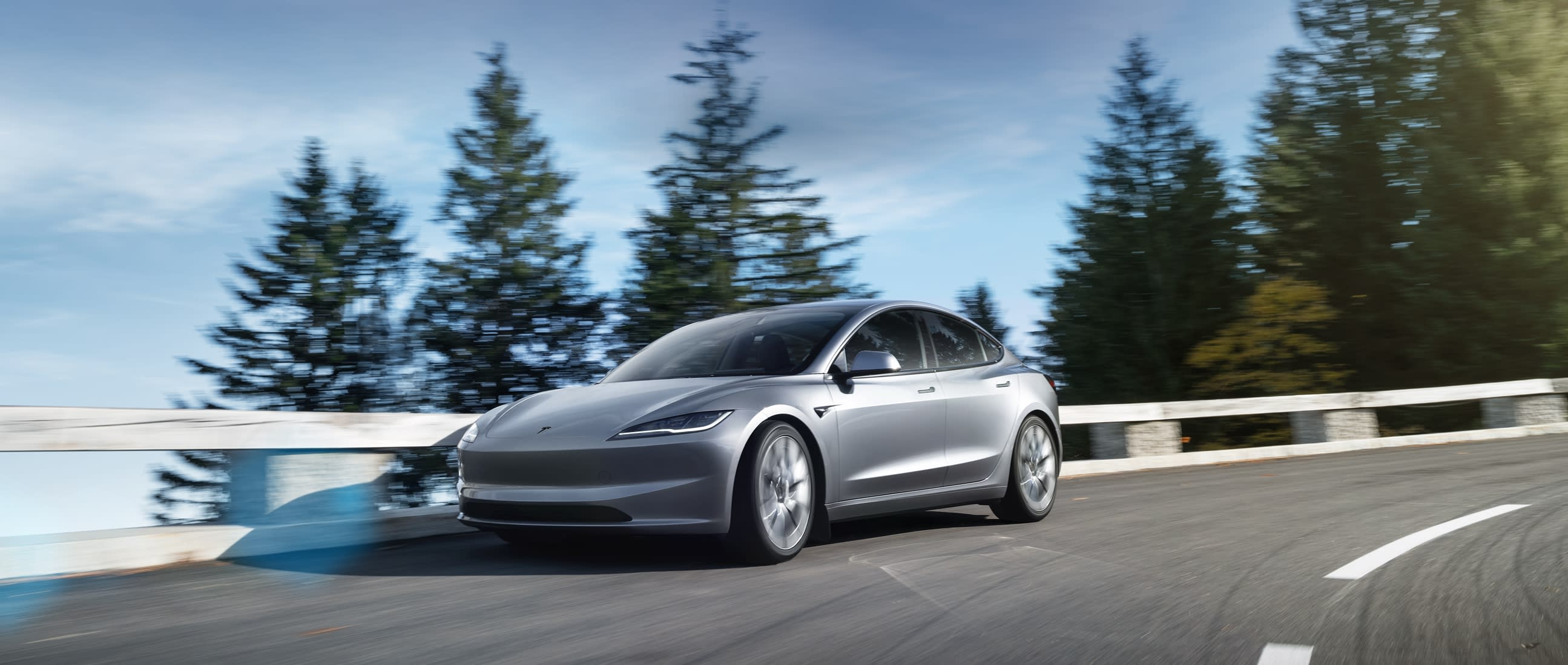 Silver Tesla Model 3 Premium sedan driving around a curved mountain road lined with pine trees
