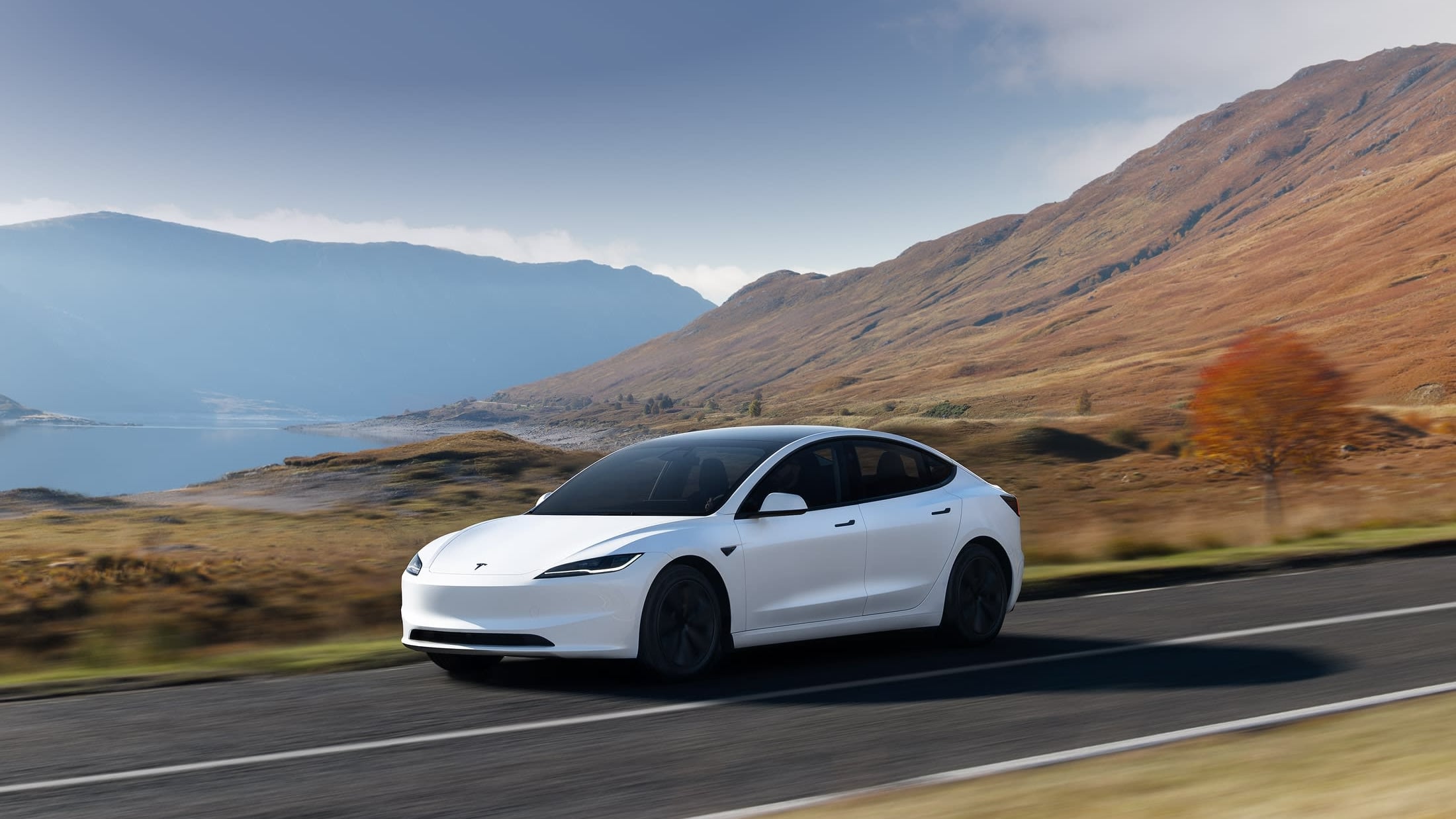 White Tesla Model 3 sedan driving along a coastal mountain road, seen from the front three-quarter angle