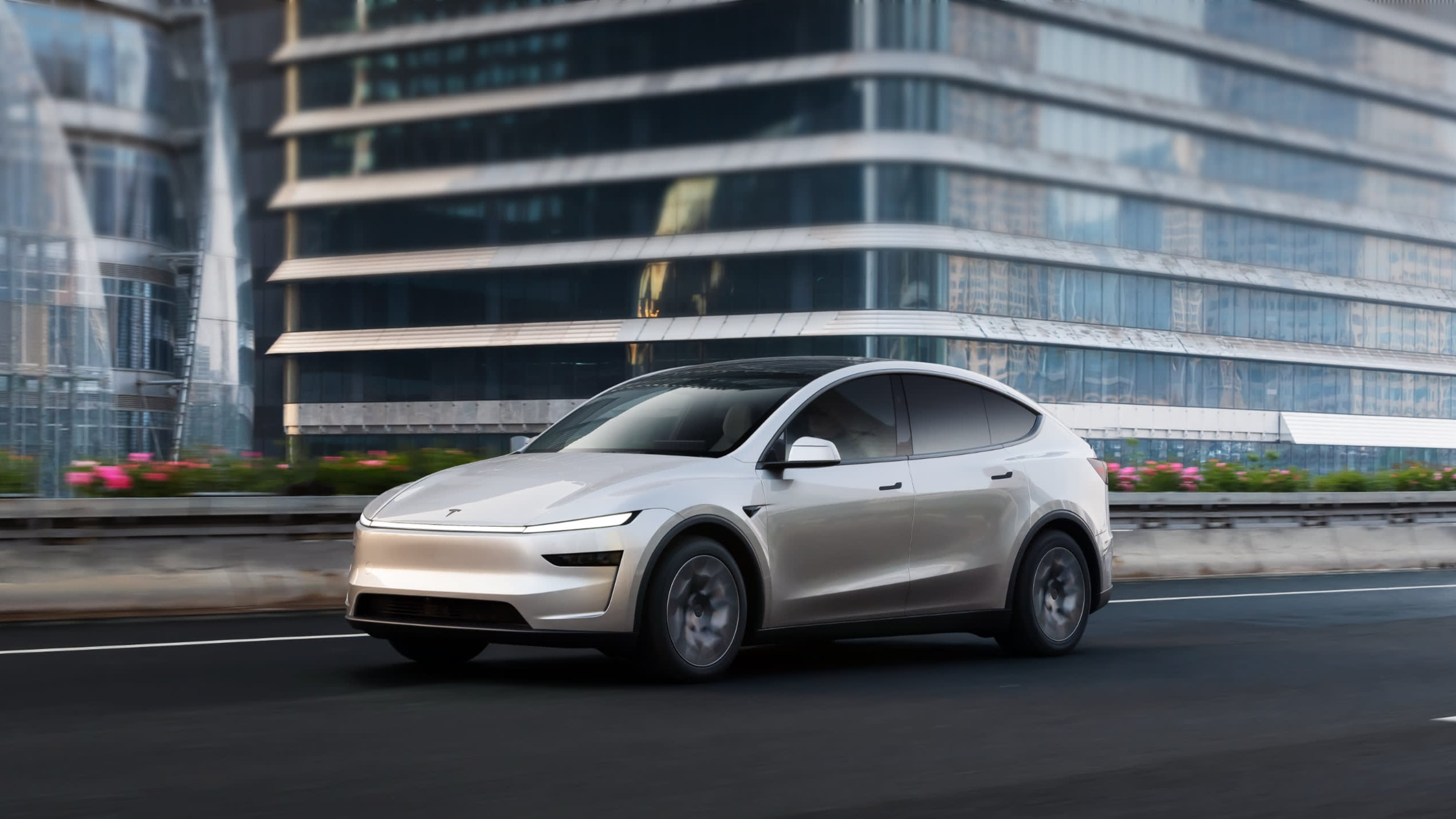 Silver Tesla Model Y L shown in front three-quarter view driving past glass office towers on an urban expressway