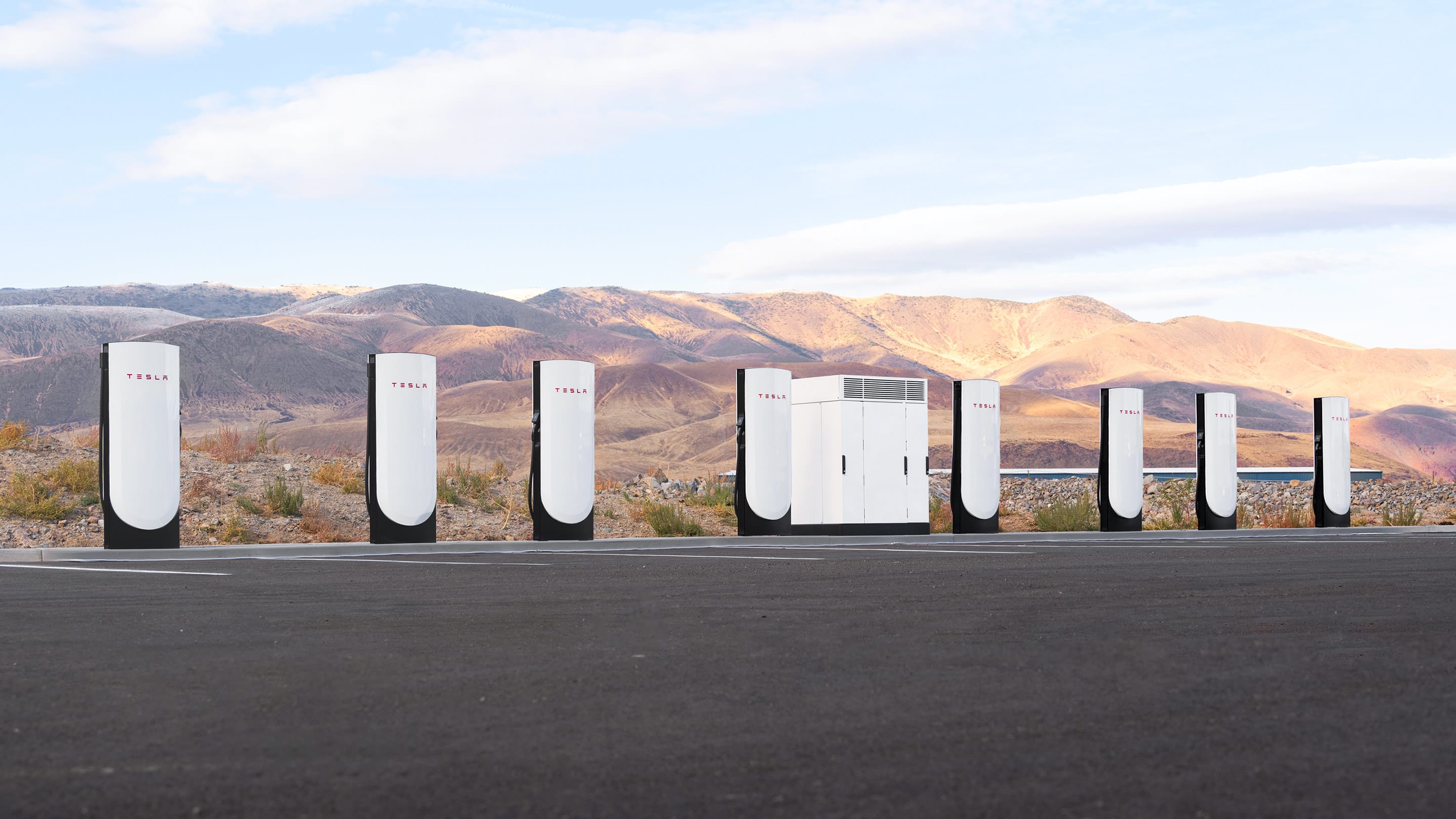 Row of white Tesla Supercharger posts and a power cabinet at an open-air charging site with hills behind
