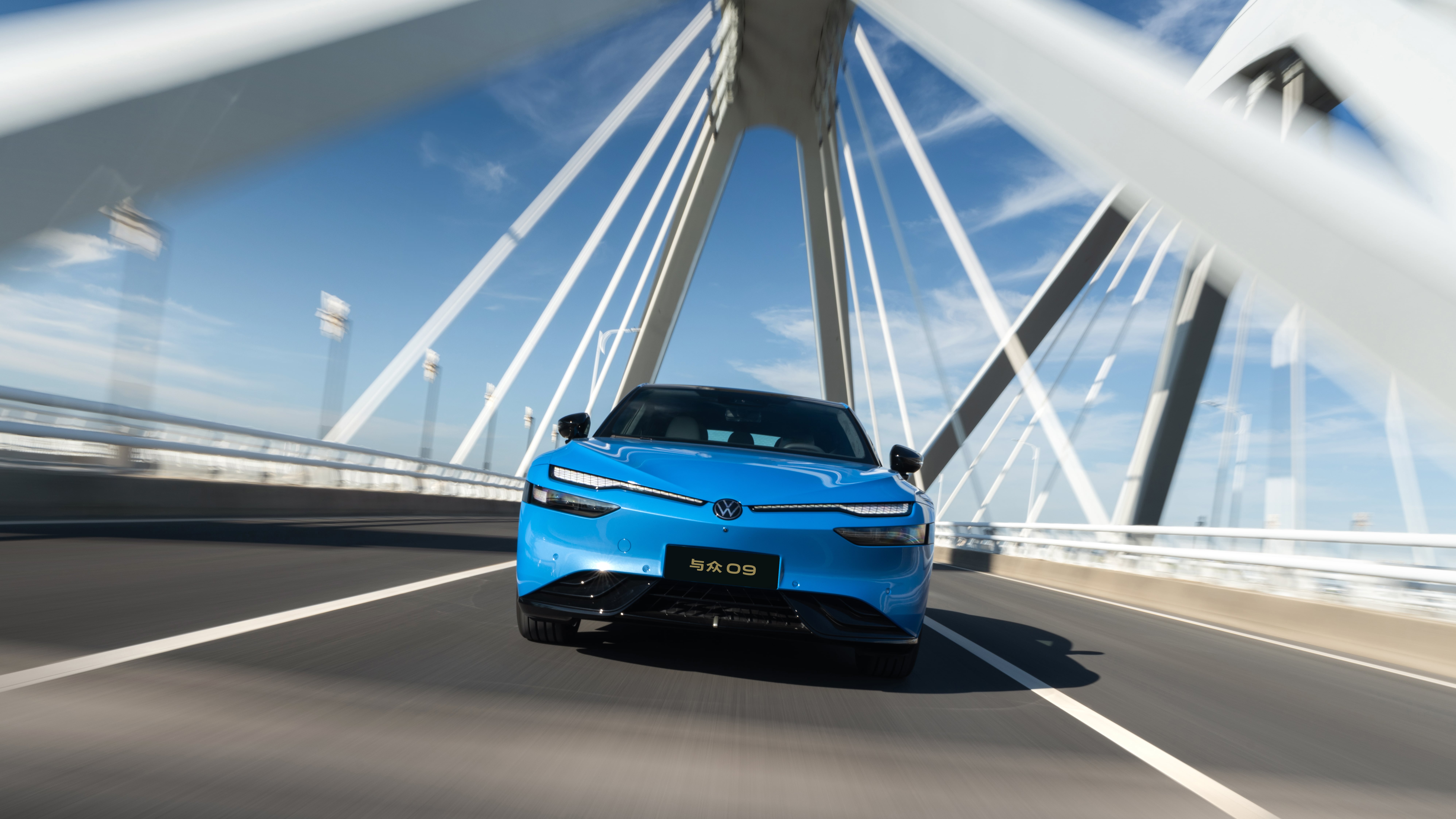 Blue Volkswagen ID. UNYX 09 fastback sedan driving head-on across a white cable-stayed bridge under a clear sky