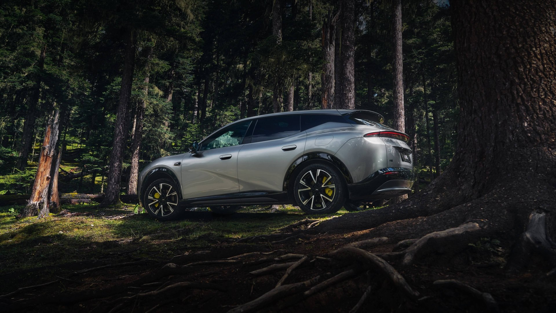 Silver XPeng Mona L05 in rear three-quarter view parked among pine trees, showing its fastback roofline and roof spoiler, official XPeng image
