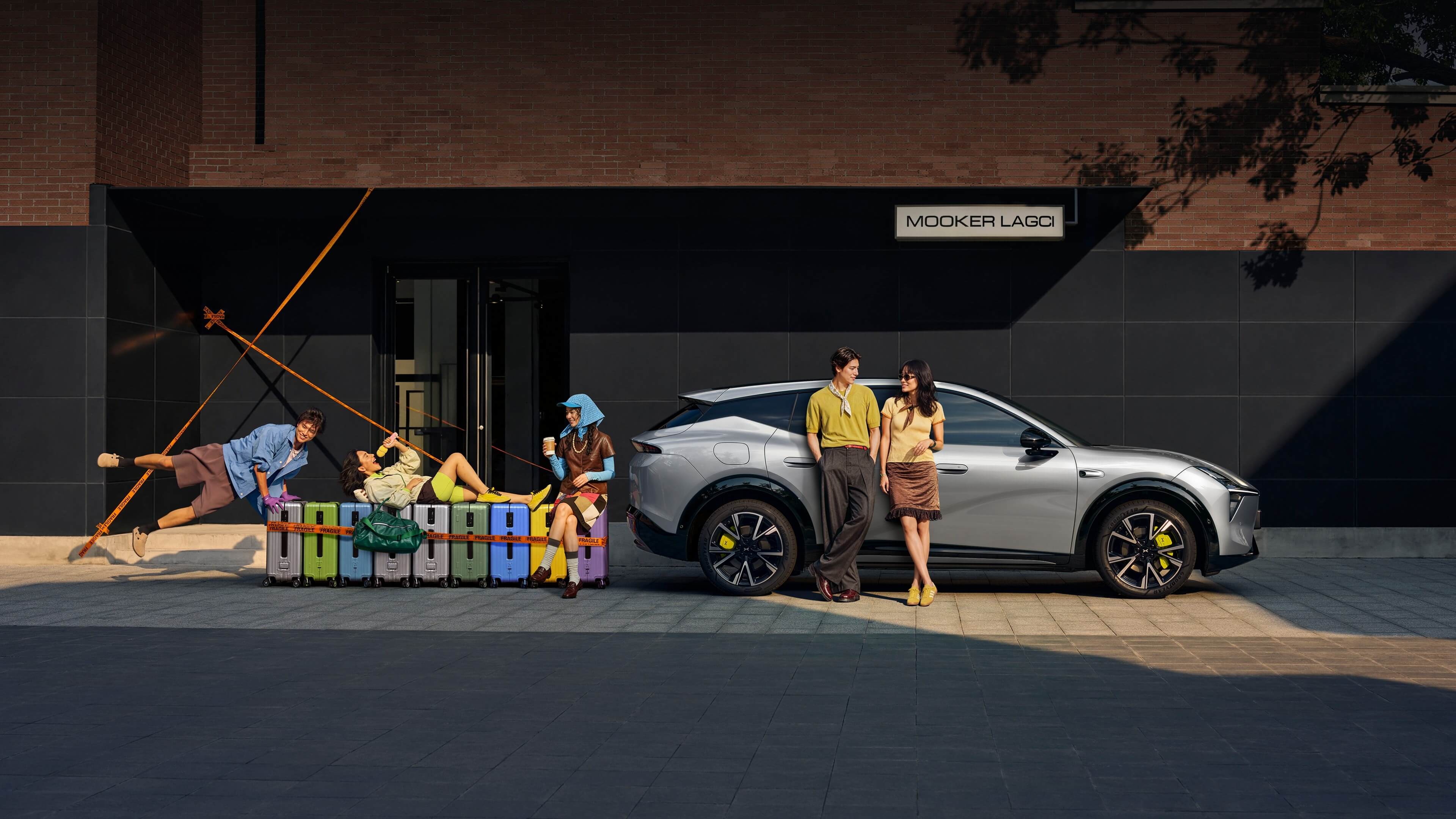 Silver XPeng Mona L05 parked on a city street beside a row of colorful suitcases, with two people standing against the car and three more posed on and around the luggage