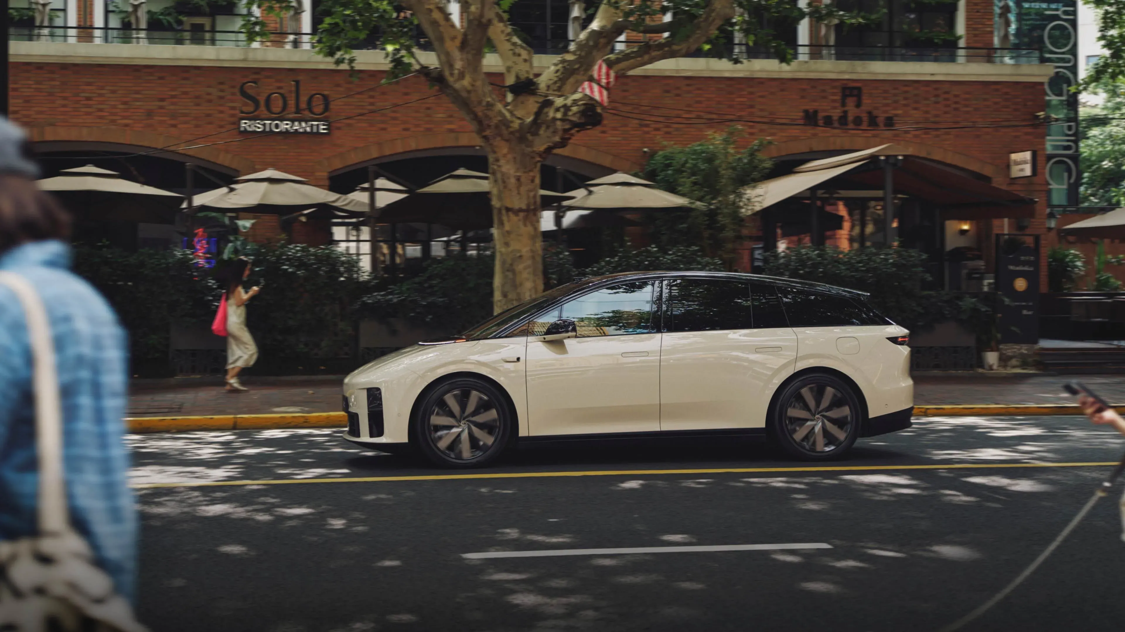 Cream-colored Li Auto i6 SUV passing a tree-lined restaurant strip on a Shanghai street